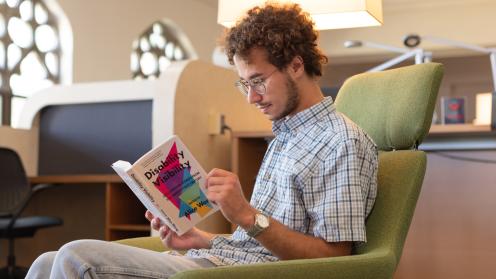KU student sitting in Watson Library on a green chair while reading "Disability Visibility." 